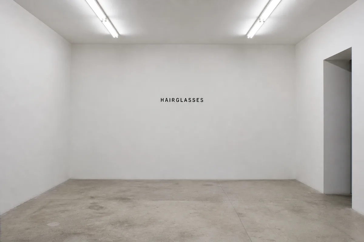 An empty white gallery room. The word Hairglasses is fixed to the far wall in small black letters. No artwork is hung.